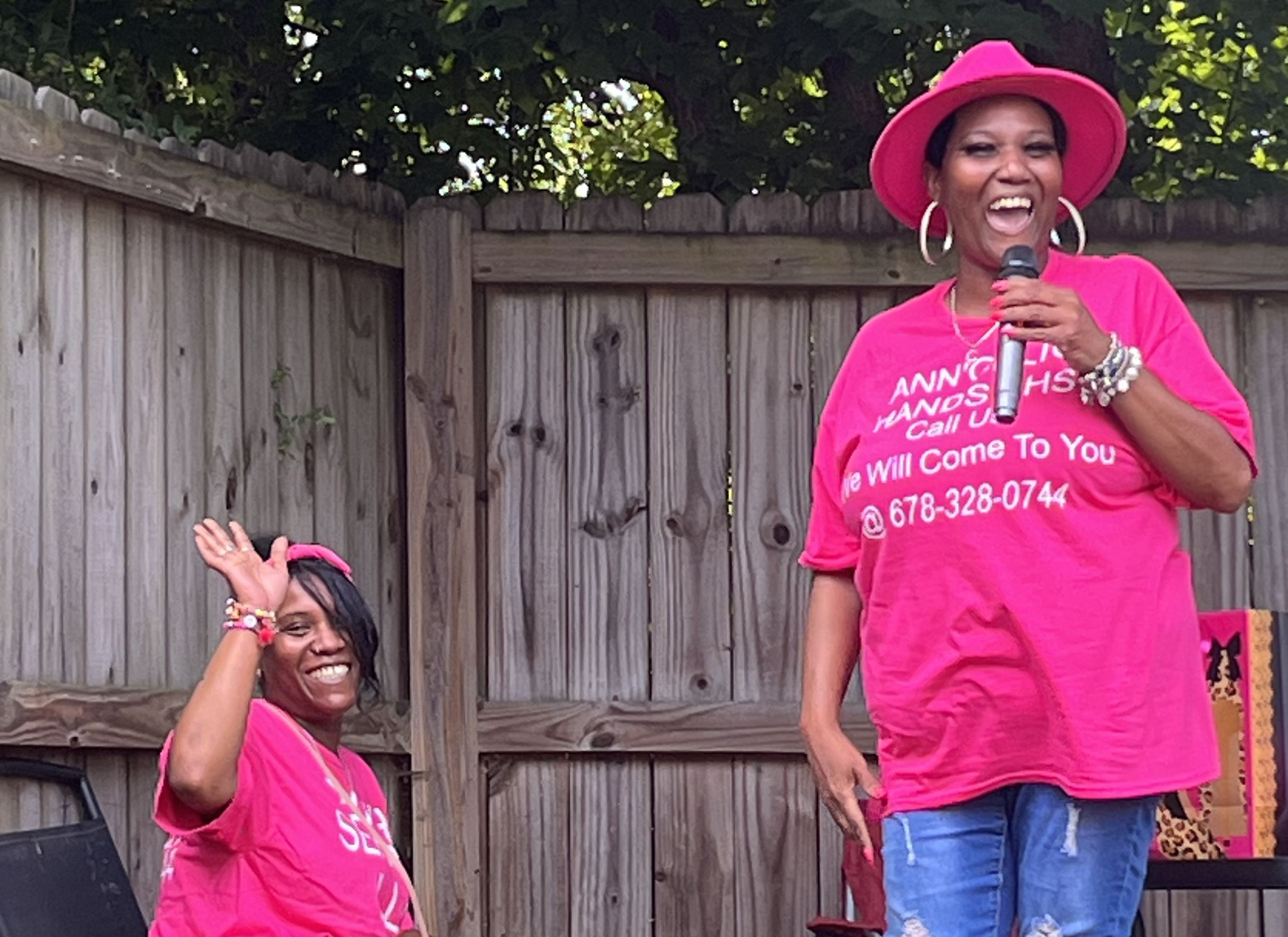 Two women of Ann'gelic Hands in pink shirts, one laughing with a microphone and one waving from a chair